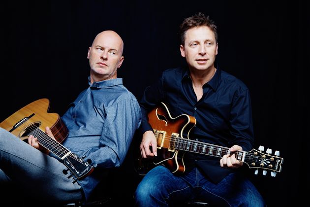 Zwei Musiker sitzen Rücken an Rücken und halten Gitarren. Der Hintergrund ist dunkel, während die Männer lässig und konzentriert wirken.