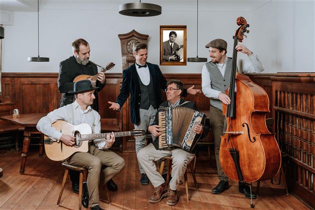 Un gruppo di cinque musicisti suona in una stanza rustica. Sono dotati di strumenti tradizionali come chitarra, contrabbasso e fisarmonica e indossano abiti eleganti.