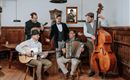 Un gruppo di cinque musicisti suona in una stanza rustica. Sono dotati di strumenti tradizionali come chitarra, contrabbasso e fisarmonica e indossano abiti eleganti.
