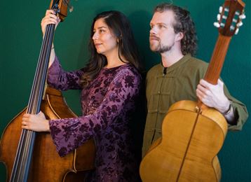 Una coppia di musicisti posa davanti a uno sfondo verde. La donna tiene un contrabbasso e l'uomo una chitarra.