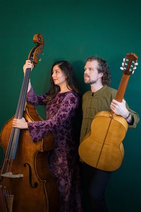 Ein Musikerpaar posiert vor einem grünen Hintergrund. Die Frau hält einen Kontrabass und der Mann eine Gitarre.