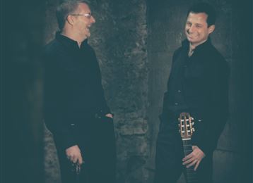 Twee mannen in zwarte kleding staan naast elkaar en glimlachen. Een van hen houdt een gitaar vast en ze lijken met elkaar te praten.