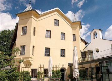 Un bâtiment élégant avec une façade jaune et des éléments historiques. Au premier plan, on peut voir des sièges et des parasols.