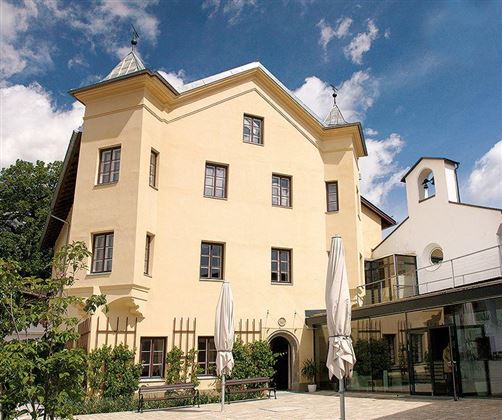 A stylish building with a yellow facade and historical elements. In the foreground, seating and umbrellas can be seen.