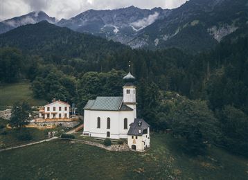 Une église pittoresque entourée de prairies vertes et de montagnes. Le ciel est nuageux, ce qui confère à la scène une atmosphère calme.