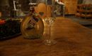 Une bouteille avec un liquide clair est posée sur une table en bois à côté d'un petit verre. À l'arrière-plan, des meubles et des plantes flous sont visibles.