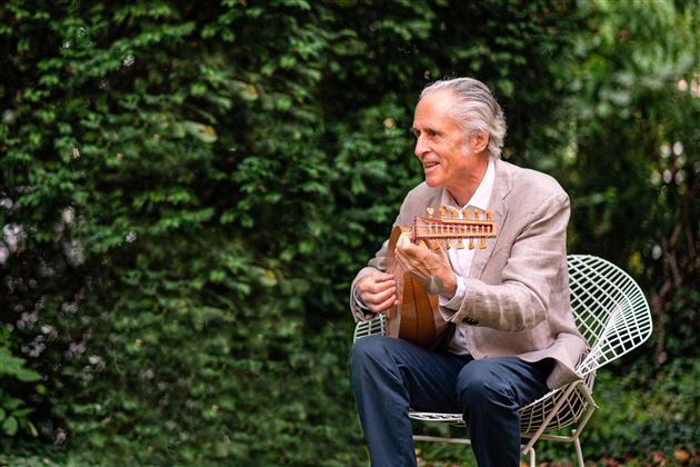 Un homme âgé est assis sur une chaise et joue d'un instrument. En arrière-plan, on peut voir des plantes vertes.