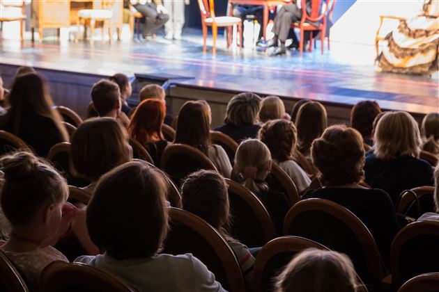 An audience is seated in a theater hall and watching the stage. The spectators are clearly visible while a performance is taking place.
