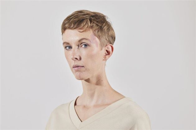 Un portrait d'une personne aux cheveux courts et châtain clair. Elle porte un haut simple beige et regarde directement l'objectif.