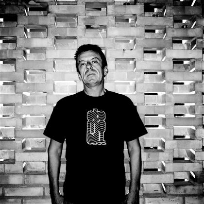 Een man staat voor een gestructureerde muur van bakstenen. Hij draagt een zwart T-shirt met een grafisch ontwerp.