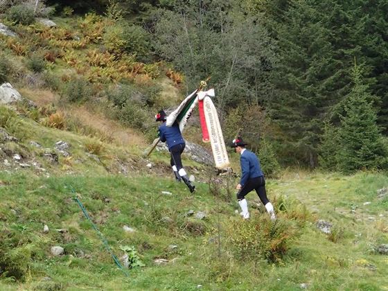 Two men are walking with a flagpole across a green meadow in the woods. The surroundings are colorful with bushes and trees.