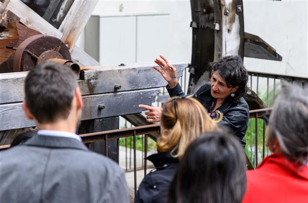 Eine Frau erklärt etwas im Freien und zeigt dabei auf eine große Maschinenstruktur. Eine Gruppe von Menschen hört aufmerksam zu.