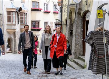 Un groupe de personnes se promène tranquillement dans une rue historique. En arrière-plan, on peut voir des fenêtres colorées et un portant à vêtements.