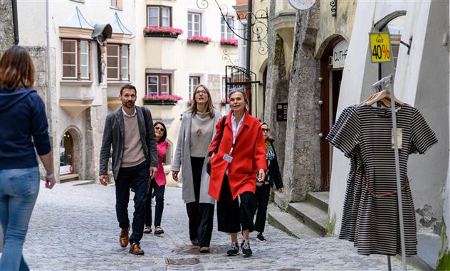 Un gruppo di persone passeggia tranquillamente attraverso una via storica. Sullo sfondo si vedono finestre colorate e un appendiabiti.