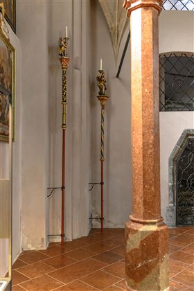 Zwei kunstvoll verzierte Kerzenständer stehen an einer Wand in einem Kircheninneren. Der Fußboden ist mit braunen Fliesen ausgelegt und ein dekorativer Säulenstütze ist zu sehen.