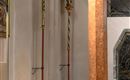 Zwei kunstvoll verzierte Kerzenständer stehen an einer Wand in einem Kircheninneren. Der Fußboden ist mit braunen Fliesen ausgelegt und ein dekorativer Säulenstütze ist zu sehen.