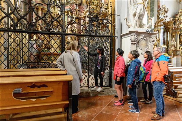 Eine Gruppe von Touristen steht vor einem kunstvollen Gitter in einer Kirche. Sie hören aufmerksam einer Führungsperson zu, die Informationen über die Kunstwerke gibt.