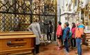 Eine Gruppe von Touristen steht vor einem kunstvollen Gitter in einer Kirche. Sie hören aufmerksam einer Führungsperson zu, die Informationen über die Kunstwerke gibt.