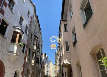 Une rue étroite avec des bâtiments colorés et un ciel bleu clair. Un panneau décoratif pend au-dessus de la rue.