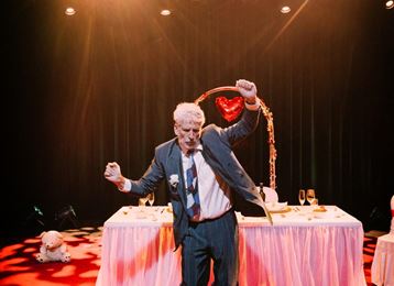 Een man in een pak danst op een podium. Op de achtergrond zijn een feestelijk gedekte tafel en een hartvormige decoratie te zien.
