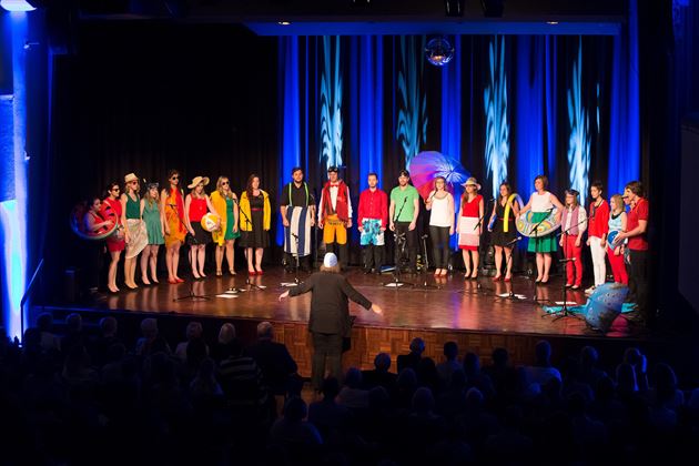Un groupe de chanteurs se tient sur scène et se produisent. Ils portent des vêtements colorés et sont entourés d'un éclairage festif.