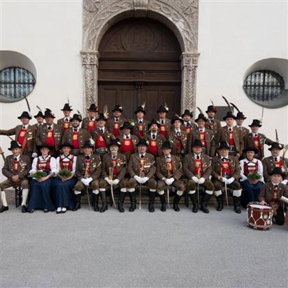 Eine Gruppe von Personen in traditioneller Tracht steht vor einer großen Tür. Sie tragen Hüte und Medaillen und halten einige Auszeichnungen in den Händen.