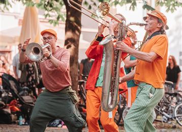 Trois musiciens jouent joyeusement dans la rue. Ils portent des vêtements colorés et sont entourés d'une atmosphère animée.