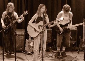 A band is performing on stage. The musicians are playing guitar and bass in a dimly lit room with colored light.