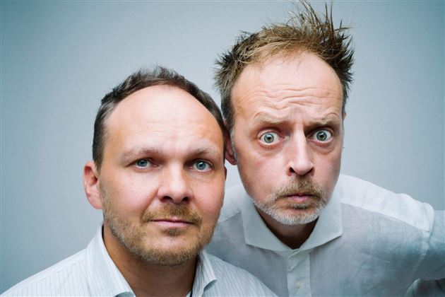 Due uomini stanno in piedi uno accanto all'altro e guardano direttamente in camera. Uno ha un'espressione sorpresa, mentre l'altro appare neutro.
