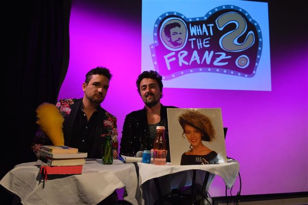 Two men are sitting at a table with eye-catching decoration. In the background, there is a colorful logo with the inscription "What the Franz."