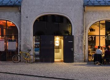 Ein angesagtes Café mit großen Fenstern in einem historischen Gebäude. Draußen sitzen Gäste an Tischen und genießen die Atmosphäre am Abend.