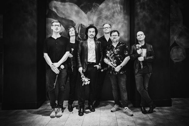 Due uomini siedono su una panchina vestiti di scuro. Indossano occhiali da sole e appaiono rilassati in un ambiente urbano.