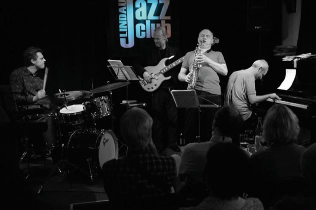 Una band jazzistica si esibisce su un palco. Suonano un pianista, un sassofonista e altri due musicisti.