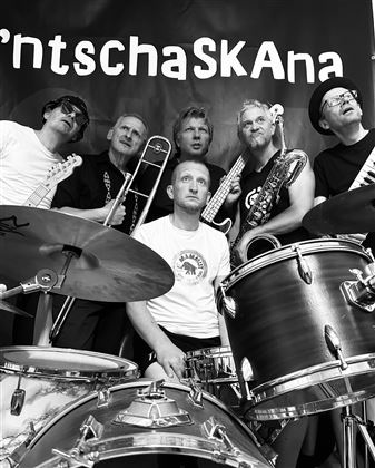 Un groupe pose devant un fond noir portant le nom "ntschaSKAna". Les musiciens se tiennent ensemble avec leurs instruments et rayonnent de joie.
