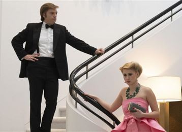 Ein Mann in einem schwarzen Anzug steht an der Treppe, während eine Frau in einem eleganten, rosa Kleid sitzt. Der Raum ist modern und hell mit einem minimalistischen Design.