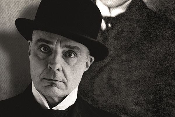 A man in a black top hat and suit is looking directly at the camera. In the background, a blurred silhouette is visible.