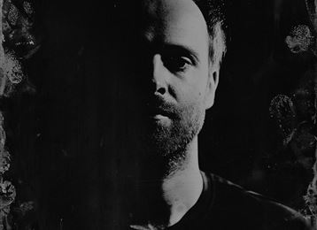A monochrome portrait of a person with a partially lit face. The background is dark and blurry, which draws the focus to the facial features.