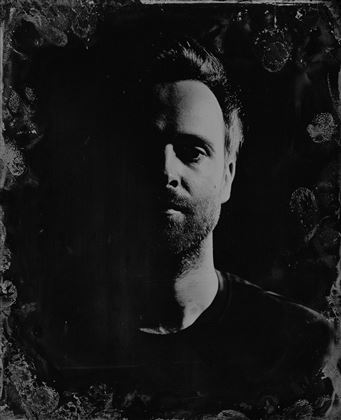 Ein monochromes Porträt einer Person mit einem teilweise beleuchteten Gesicht. Der Hintergrund ist dunkel und unscharf, was den Fokus auf die Gesichtszüge lenkt.