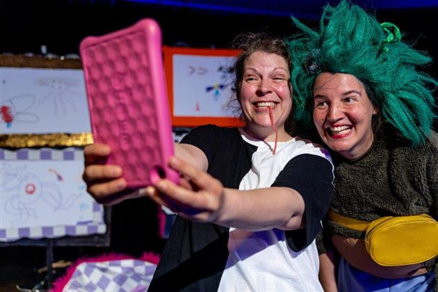 Zwei fröhliche Frauen machen ein Selfie auf einer Bühne. Im Hintergrund sind bunte Zeichnungen sichtbar.