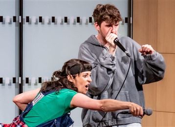 Une femme en chemise verte réalise une performance énergique, tandis qu'un jeune homme rappe avec un microphone. Tous deux semblent passionnés par la musique.