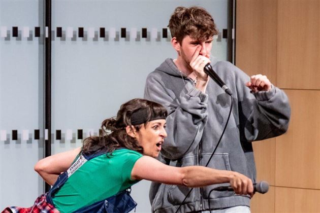 Een vrouw in een groen shirt geeft een energieke performance, terwijl een jonge man met een microfoon rapt. Beiden lijken enthousiast van de muziek te zijn.