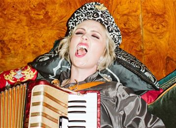 A woman wearing an animal-print hat enthusiastically plays an accordion. She is relaxed on a sofa and sings contentedly.