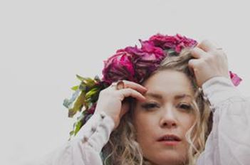 Eine Frau mit lockigem Haar trägt einen Kranz aus rosa Blumen. Ihr Gesichtsausdruck ist nachdenklich und verträumt.