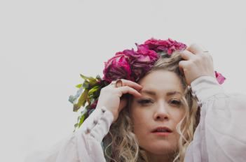 Une femme aux cheveux bouclés porte une couronne de fleurs roses. Son expression est pensive et rêveuse.