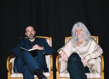 Twee mannen zitten ontspannen in stijlvolle fauteuils, een met een notitieboek in de hand. De man rechts heeft lange, zilveren haren en draagt een lichthemd.