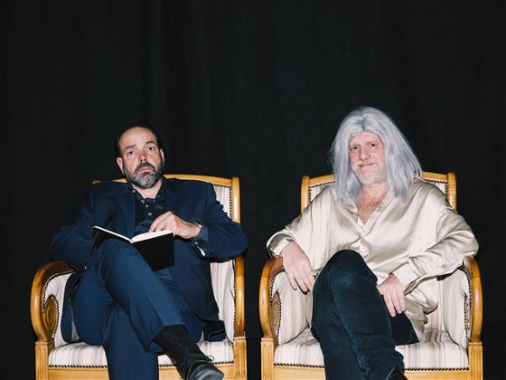 Two men are sitting relaxed in stylish armchairs, one with a notebook in his hand. The man on the right has long, silver hair and is wearing a light shirt.