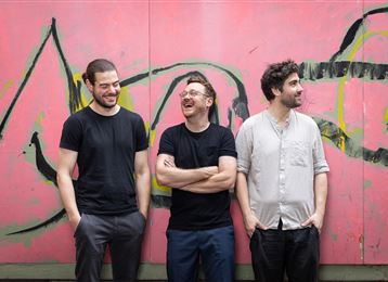 Trois hommes sourient devant un mur coloré avec des graffitis. Ils portent des vêtements décontractés et semblent détendus.