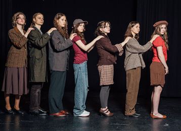 Eine Gruppe von Frauen in verschiedenen Outfits steht in einer Reihe auf einer Bühne. Sie haben unterschiedliche Posen und Gesichtsausdrücke.