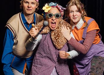 Trois personnes avec des tenues humoristiques posent joyeusement. La personne du milieu porte des lunettes voyantes et a une expression énergique.
