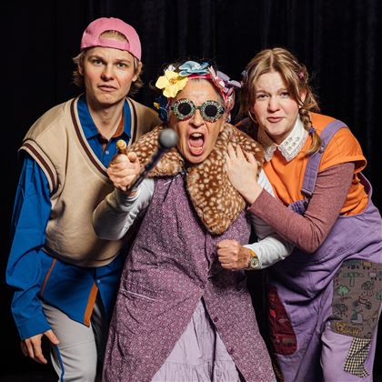 Three people in humorous outfits are happily posing. The person in the middle is wearing striking glasses and has an energetic expression.